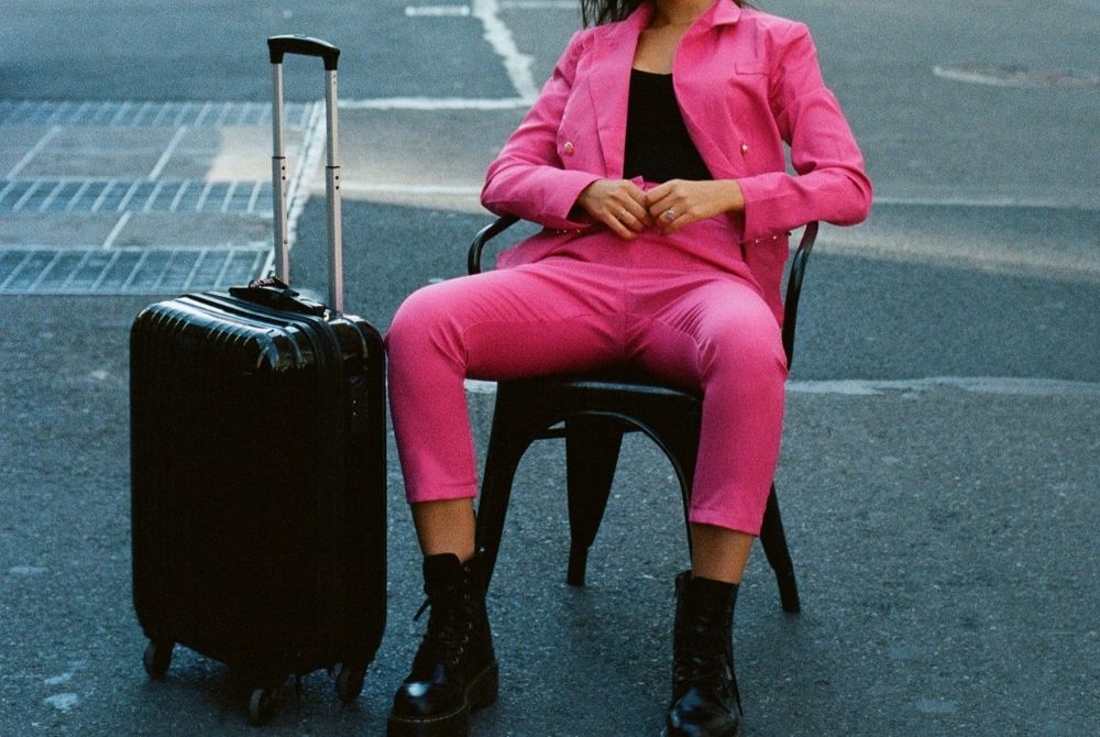 frau in pinkem hosenanzug sitzt auf stuhl neben koffer